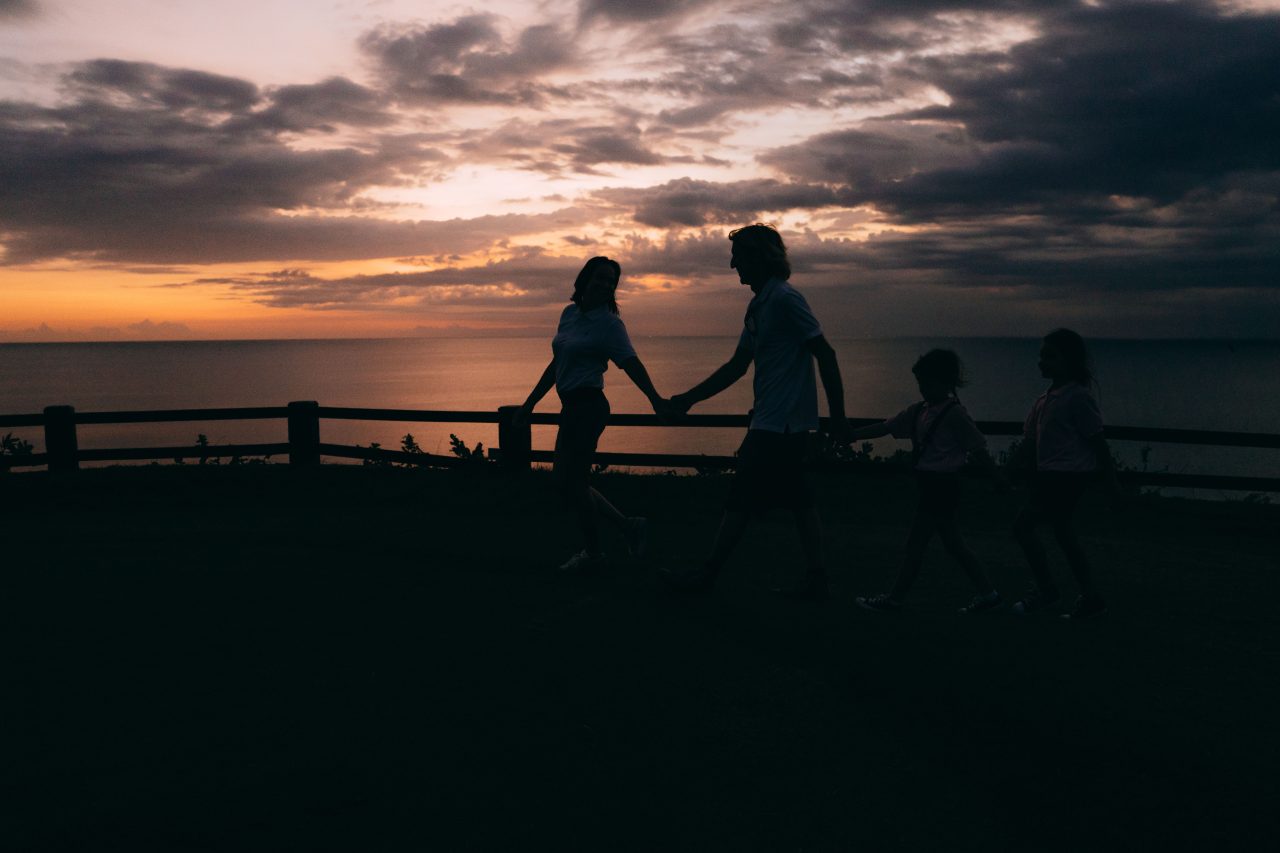 A family of four silhouetted walking hand in hand along a coastal path at sunset.